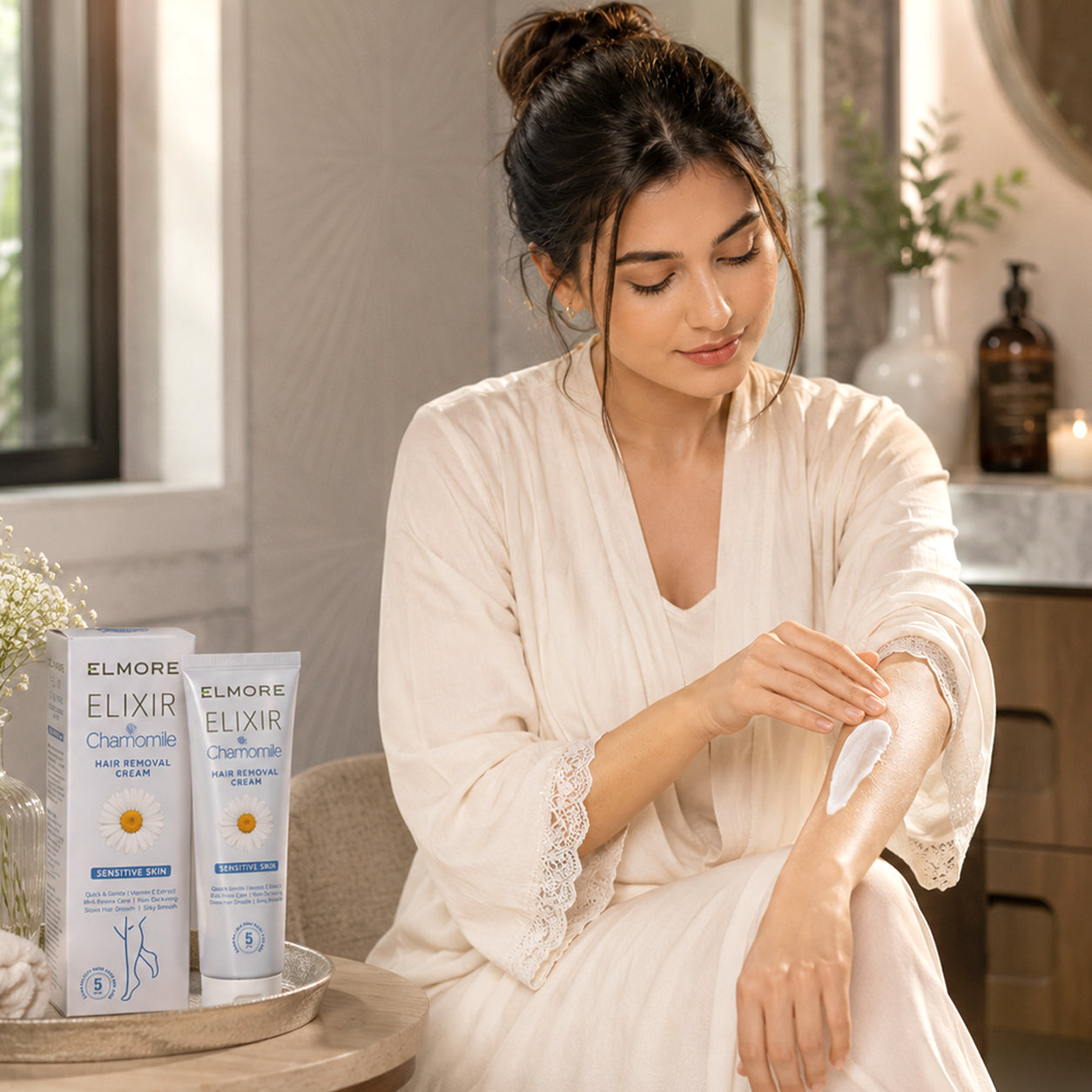 A woman in a cream robe applies cream to her arm while standing beside two boxes of ELMORE ELIXIR Chamomile hair removal cream on a small table in a softly lit bathroom setting.