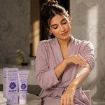 A woman in a lavender robe applies a white body cream to her upper arm while sitting in a spa-like setting. Two boxes of ELMORE ELIXIR Lavender hair removal cream are visible on the table beside her.